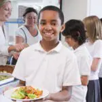 Schoolboy holding plate of lunch in school cafeteria smiling at camera
